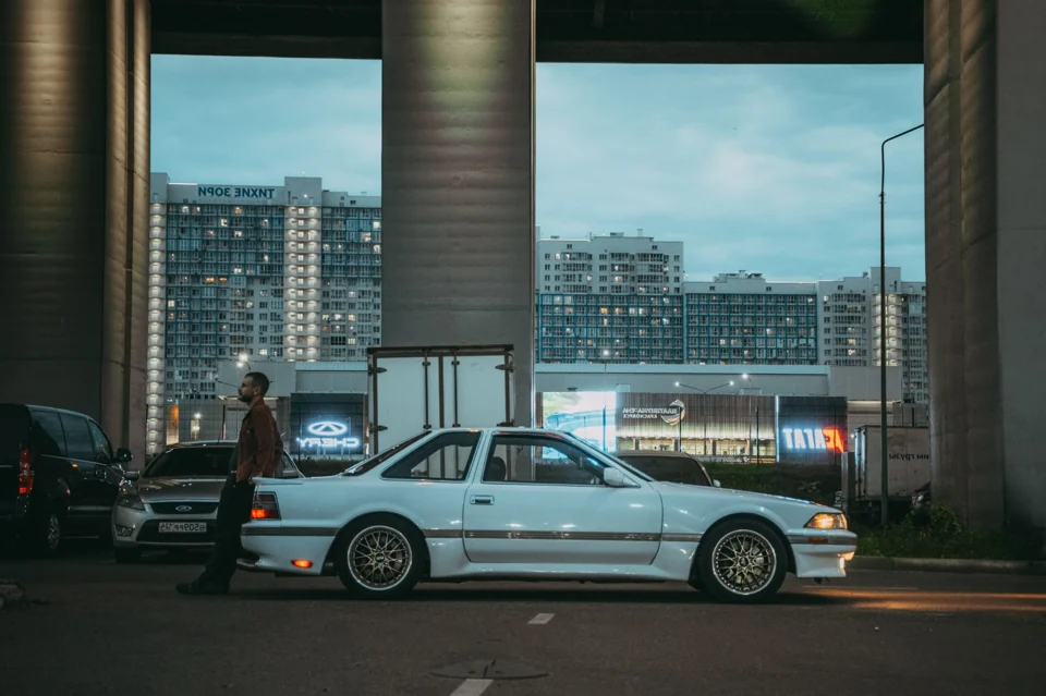 Светлый Toyota Soarer на фоне вечернего города и массивных опор эстакады.